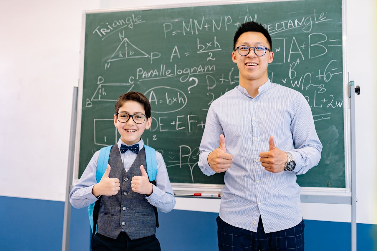 A teacher and student smiling in front of a chalkboard with mathematical formulas, both giving thumbs up.
