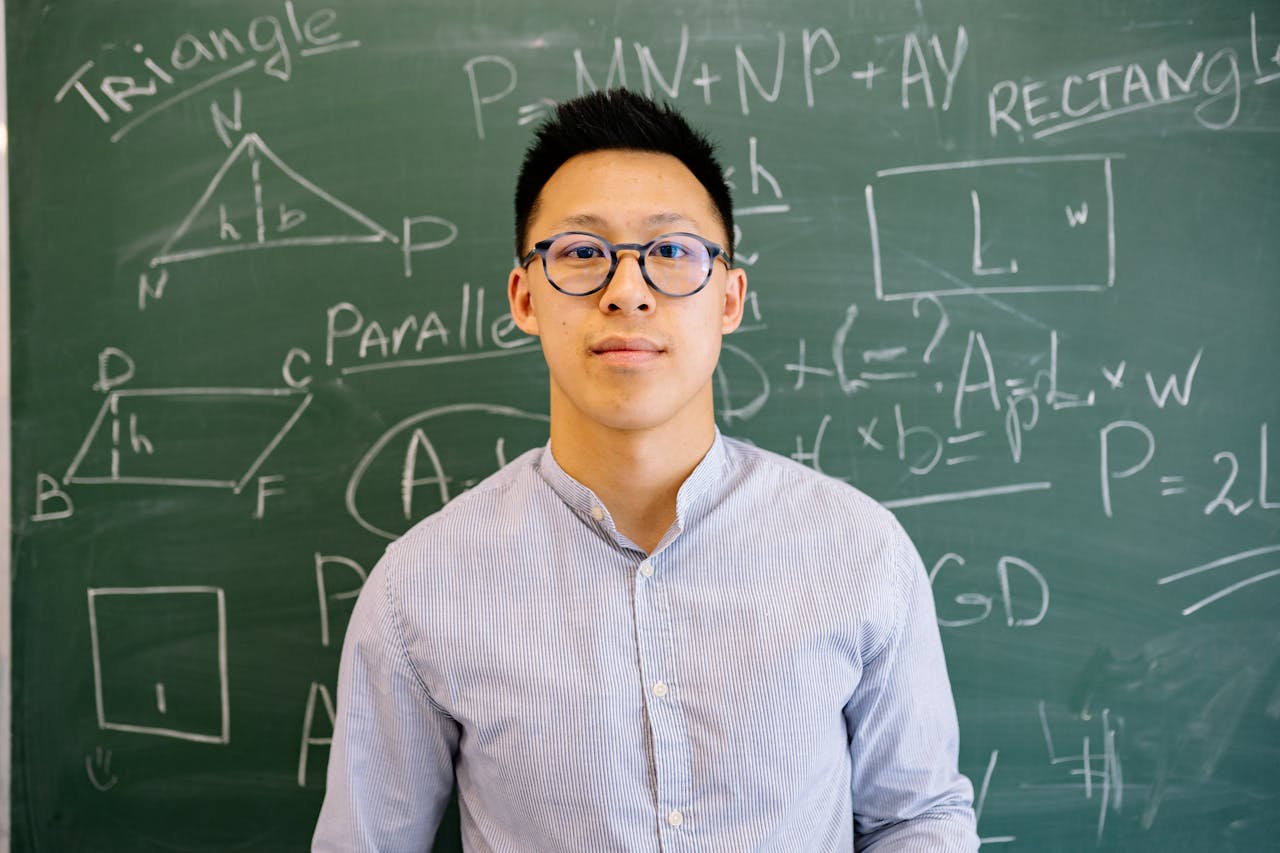 Educator standing before chalkboard filled with geometry and algebra equations, wearing glasses and long-sleeved shirt.